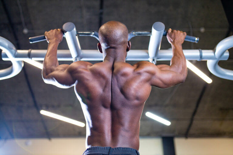 Homme effectuant des tractions en pronation à la barre fixe, vu de dos, dans une salle de sport moderne.
